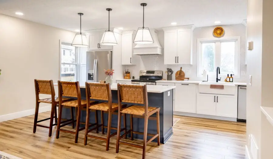 White shaker kitchen with navy blue island featuring four woven leather barstools. Bright space includes white quartz countertops, farmhouse sink, black fixtures, herringbone tile backsplash, and stainless appliances. Three pendant lights over island, hardwood flooring, and natural wood accents add warmth. Design balances classic elements with modern touches for timeless appeal.
