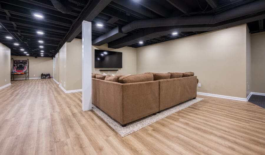 Finished basement featuring media area with tan sectional sofa and wall-mounted TV centered in cozy viewing space. Open layout includes basketball arcade game visible in the distance. Black painted exposed ceiling with recessed lighting maintains height while concealing utilities and ductwork. Light vinyl plank flooring unifies the space, with beige walls and white trim creating a bright, inviting atmosphere. Strategic white column defines spaces while allowing for flow between entertainment zones.